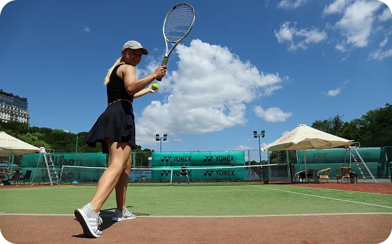 female tennis player preparing to hit the ball on an outdoor court under blue sky with clouds, showing athletic stance, focus, and active tennis gameplay