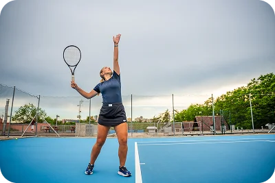 female tennis player performing a serve on a blue outdoor court under cloudy sky, showing athletic motion, focus, and competitive tennis training