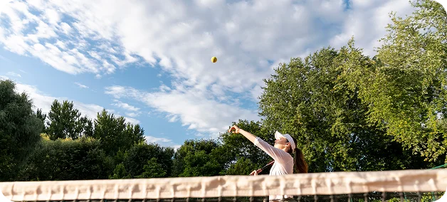 female tennis player serving the ball on an outdoor court during a sunny day with blue sky and trees, capturing motion, focus, and active sports lifestyle